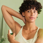 Woman raising one arm and applying Weleda deodorant spray to her underarm in front of a mirror.