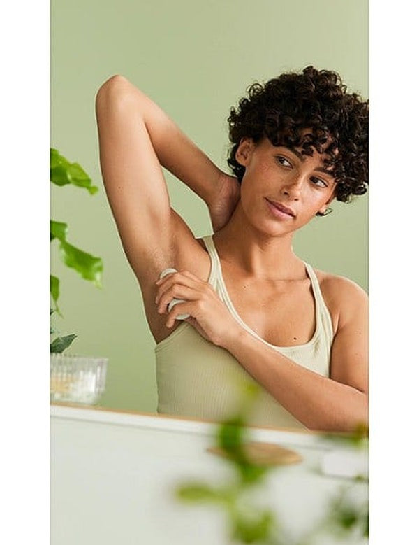 Woman applying Weleda Citrus 24h Roll-On Deodorant beneath her arm in a bright green bathroom setting.