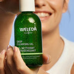 Smiling woman holding a green bottle of Weleda Deep Cleansing Gel 150ml against a soft blue background.