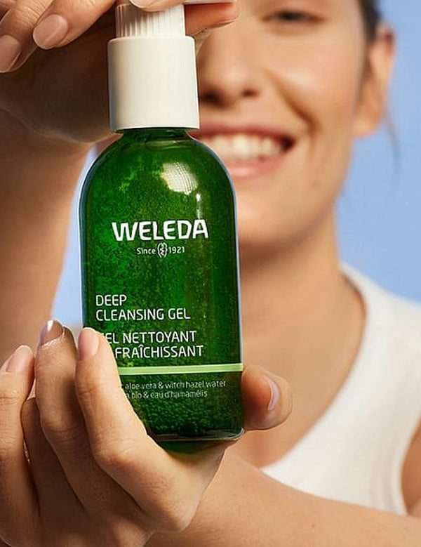 Smiling woman holding a green bottle of Weleda Deep Cleansing Gel 150ml against a soft blue background.