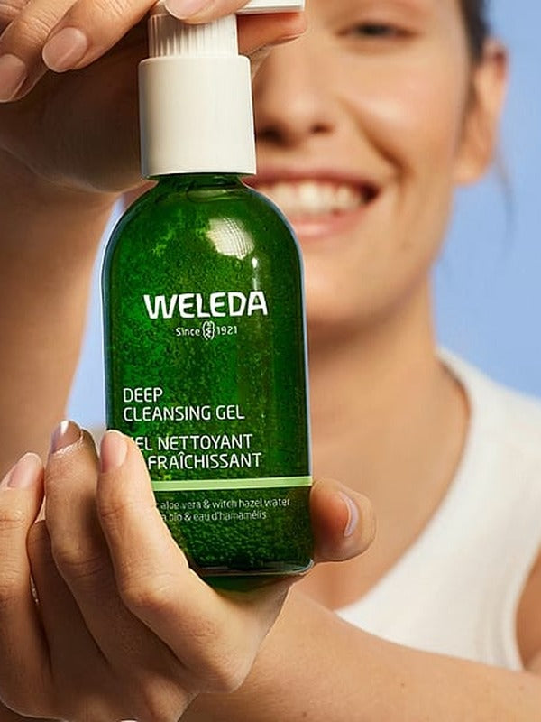 Smiling woman holding a green bottle of Weleda Deep Cleansing Gel 150ml against a soft blue background.