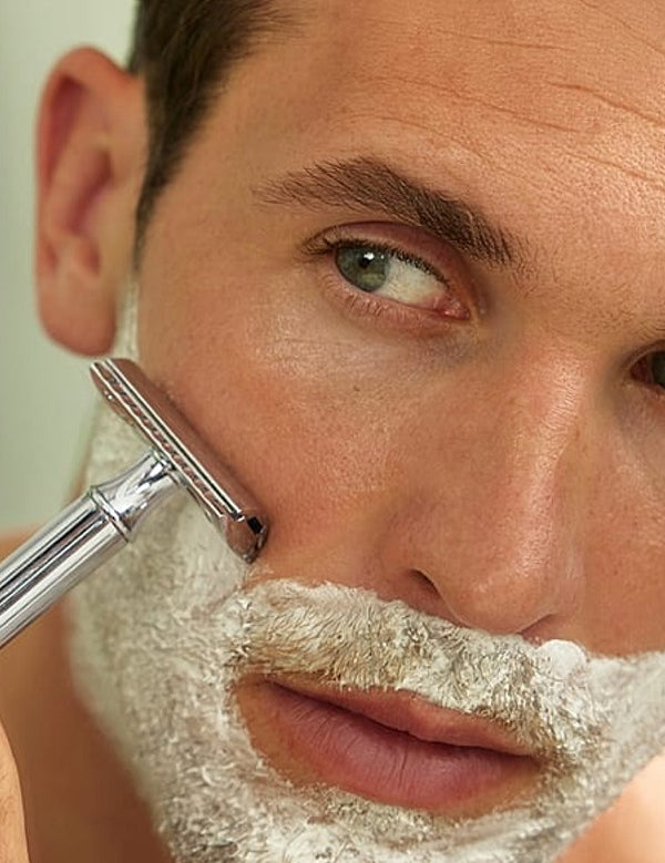 Man shaving in a mirror with Weleda For Men Shaving Cream lathered across his face.