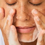 Dark skin woman with short curly hair with her eyes closed washing her face with a face cleansing foam.