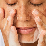 Dark skin woman with short curly hair with her eyes closed washing her face with a face cleansing foam.