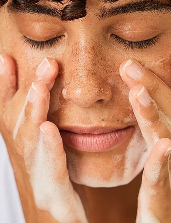 Dark skin woman with short curly hair with her eyes closed washing her face with a face cleansing foam.