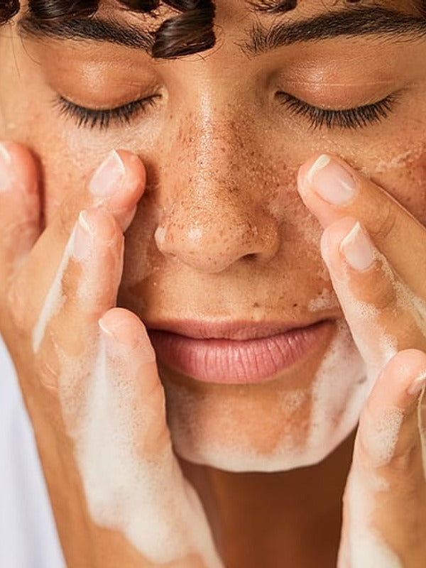 Dark skin woman with short curly hair with her eyes closed washing her face with a face cleansing foam.