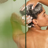 Woman washing and lathering her hair in the shower, photographed through green-tinted glass with trailing plants.