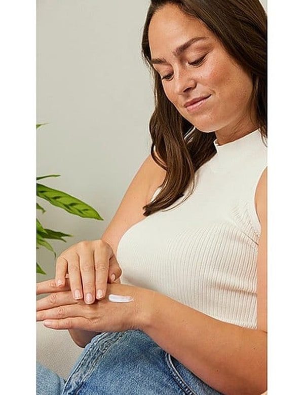 Woman smoothing a small amount of white Weleda hand cream over the back of her hand in a bright, leafy setting.