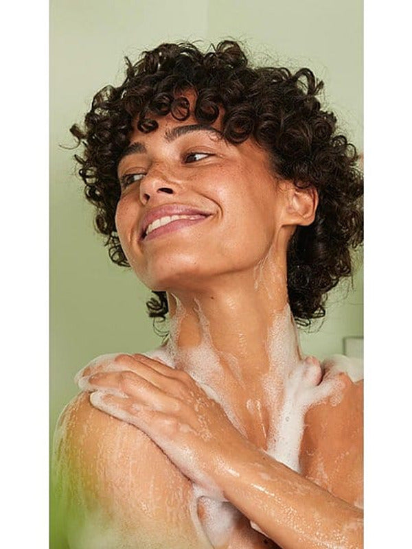 Woman smiling while applying Weleda Happiness Grapefruit Shower Gel across her shoulder in the shower.