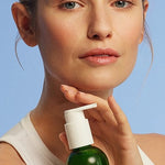 Young woman holding a bottle of Make-up Removal Cleansing Oil on a light blue background.