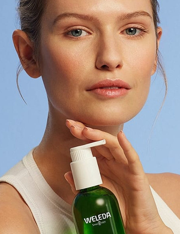 Young woman holding a bottle of Make-up Removal Cleansing Oil on a light blue background.