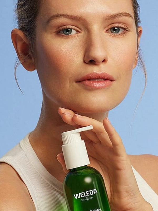 Young woman holding a bottle of Make-up Removal Cleansing Oil on a light blue background.