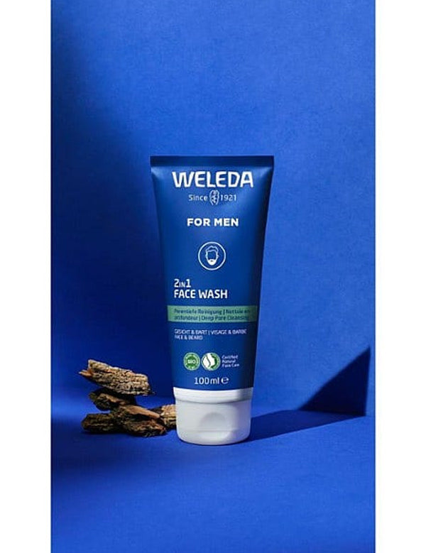 Weleda Men 2 in 1 Face Wash tube standing upright with pieces of liquorice root against a rich blue studio background.