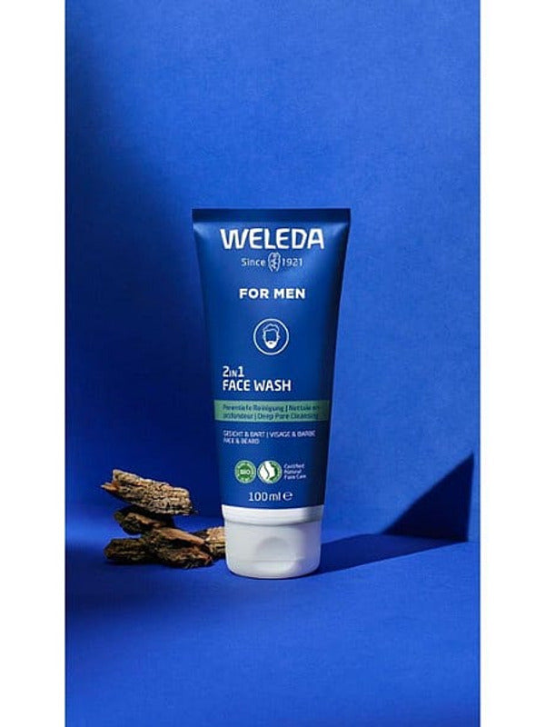 Weleda Men 2 in 1 Face Wash tube standing upright with pieces of liquorice root against a rich blue studio background.