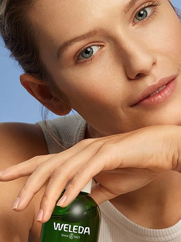 Young woman with clear skin posing with Weleda Refining Toner against a soft blue studio background.
