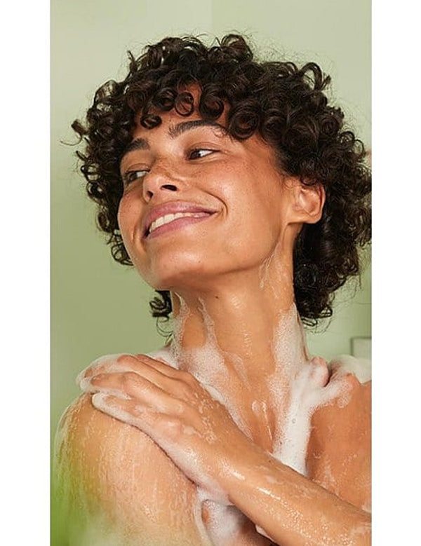 Woman smiling while applying Weleda Relax Lavender Shower Cream across her shoulder in the shower.
