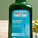 Revitalising Hair Tonic bottle on a sage green background with its ingredients rosemary leaves and horseradish root.