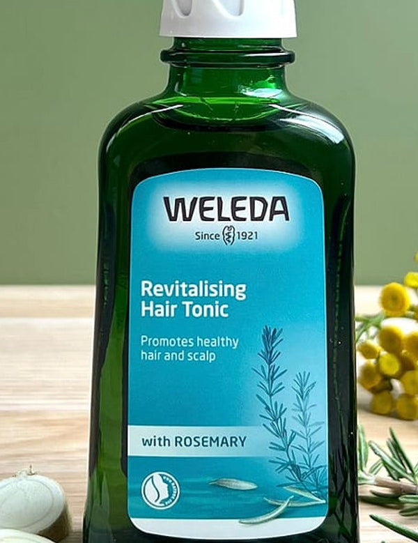 Revitalising Hair Tonic bottle on a sage green background with its ingredients rosemary leaves and horseradish root.