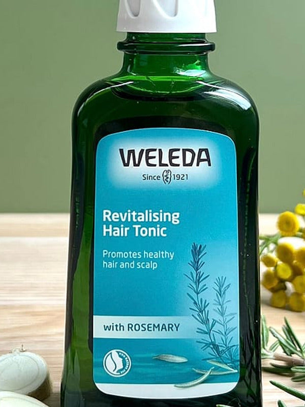 Revitalising Hair Tonic bottle on a sage green background with its ingredients rosemary leaves and horseradish root.