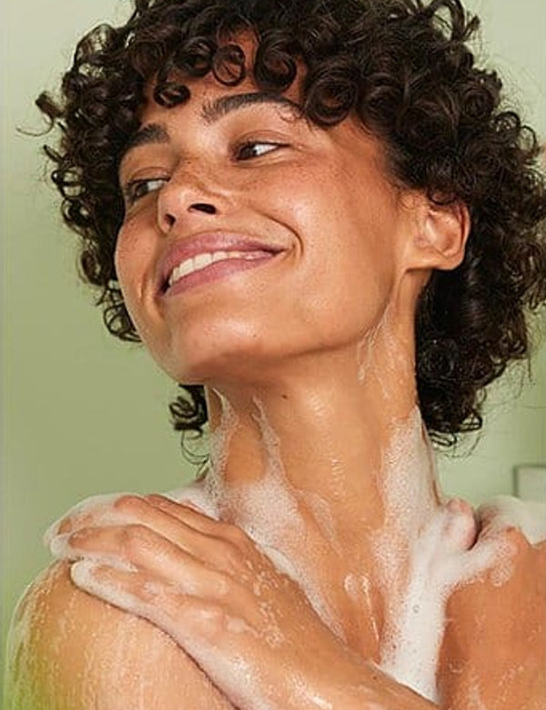 Smiling woman lathering Weleda Vitality Sea Buckthorn Shower Cream over her shoulders and neck.