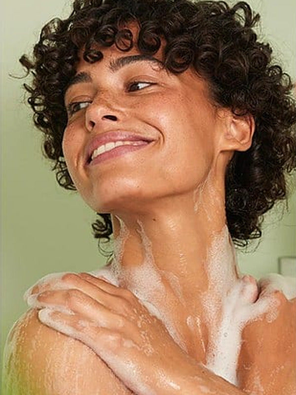 Smiling woman lathering Weleda Vitality Sea Buckthorn Shower Cream over her shoulders and neck.