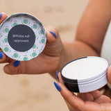 Hands holding an open tin of WooHoo Tux deodorant paste