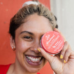Smiling woman holding WooHoo Urban deodorant paste beside her face against a red background