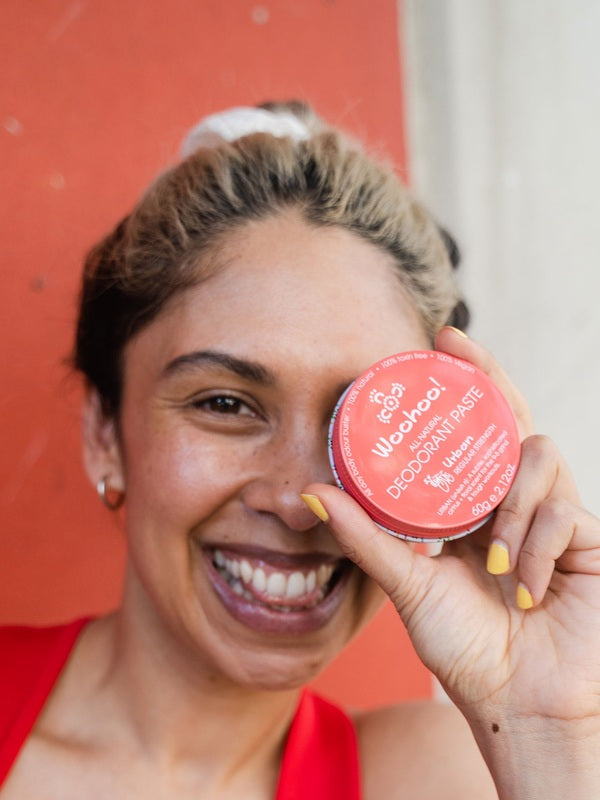 Smiling woman holding WooHoo Urban deodorant paste beside her face against a red background