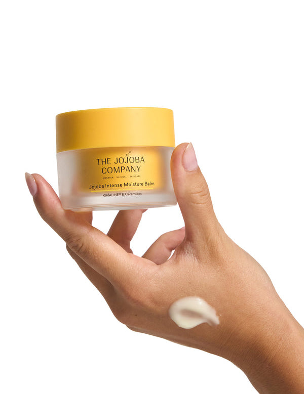 Hand holding a jar of The Jojoba Company Jojoba Intense Moisture Balm with a smear of balm