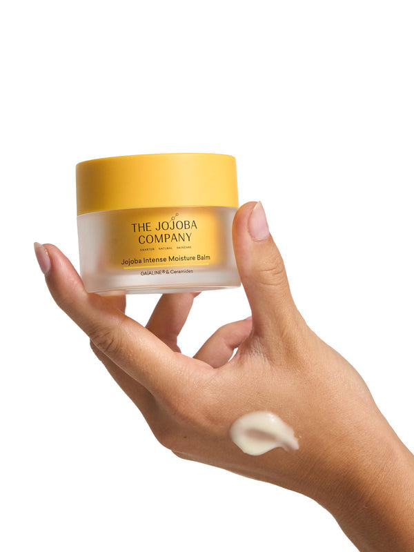 Hand holding a jar of The Jojoba Company Jojoba Intense Moisture Balm with a smear of balm