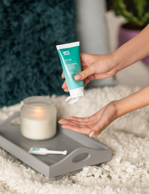 Hands dispensing YES WB Water-Based Lubricant beside a bed