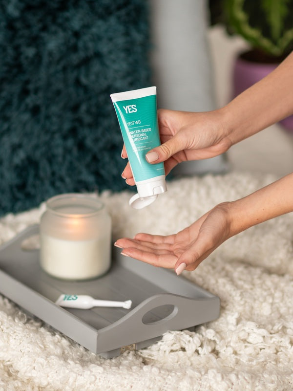 Hands dispensing YES WB Water-Based Lubricant beside a bed